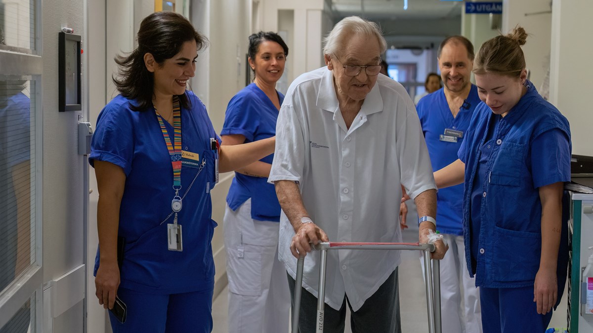 Äldre man med rullator i vita och gråa sjukhuskläder omringad av blåklädd vårdpersonal