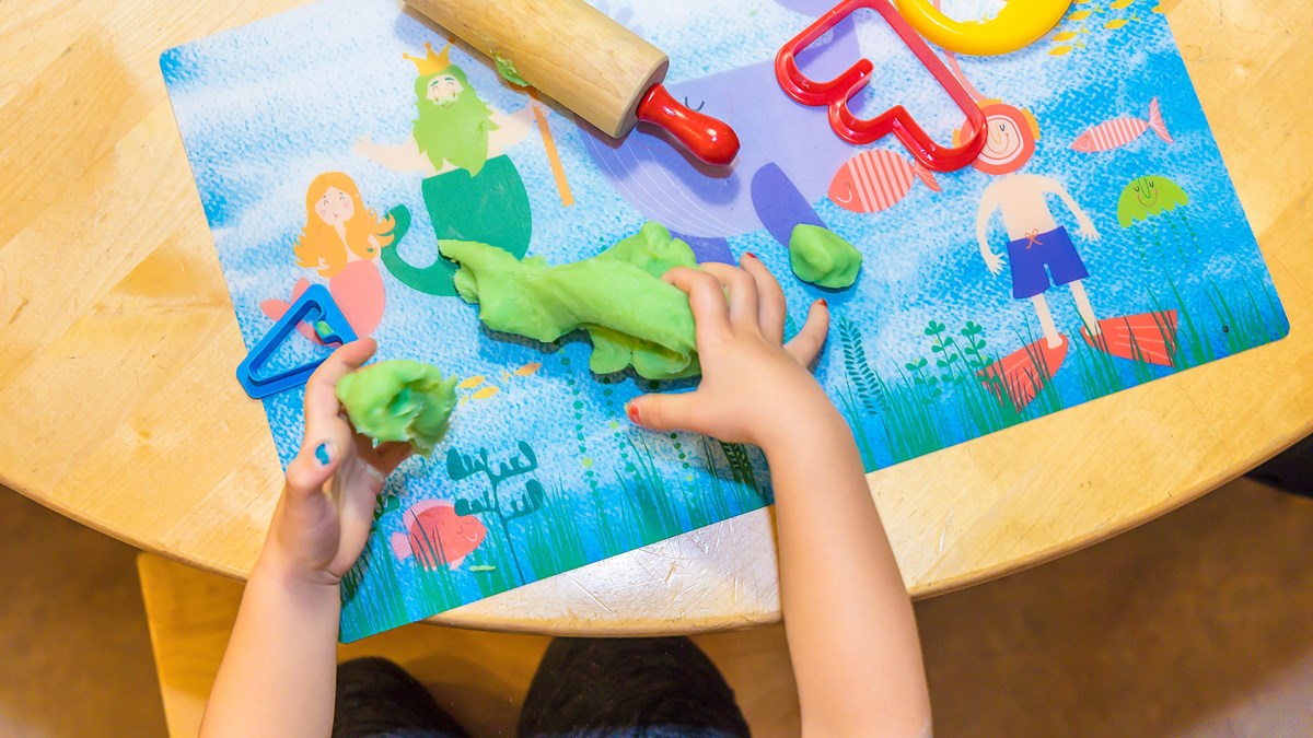 Foto taget ovanifrån på ett barn som leker med modell-lera vid ett bord. Det finns en liten kavel och några kakformar intill barnets händer.