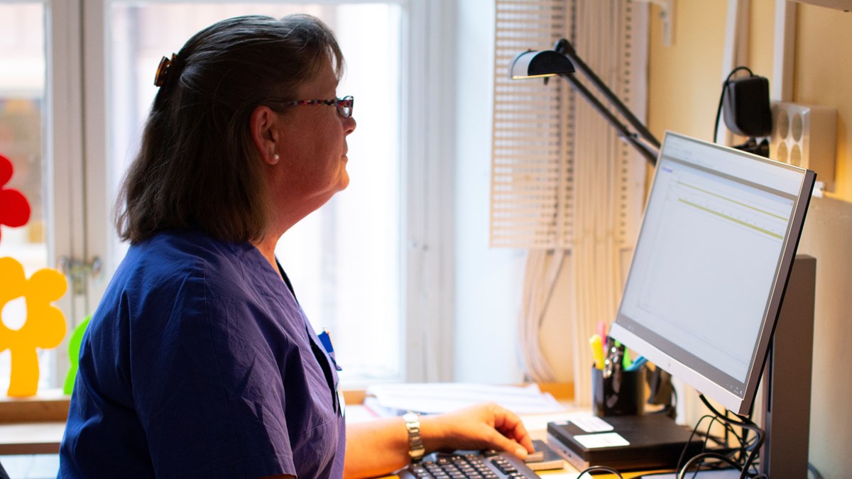 Kvinna i arbetskläder sitter vid skrivbord och dator.