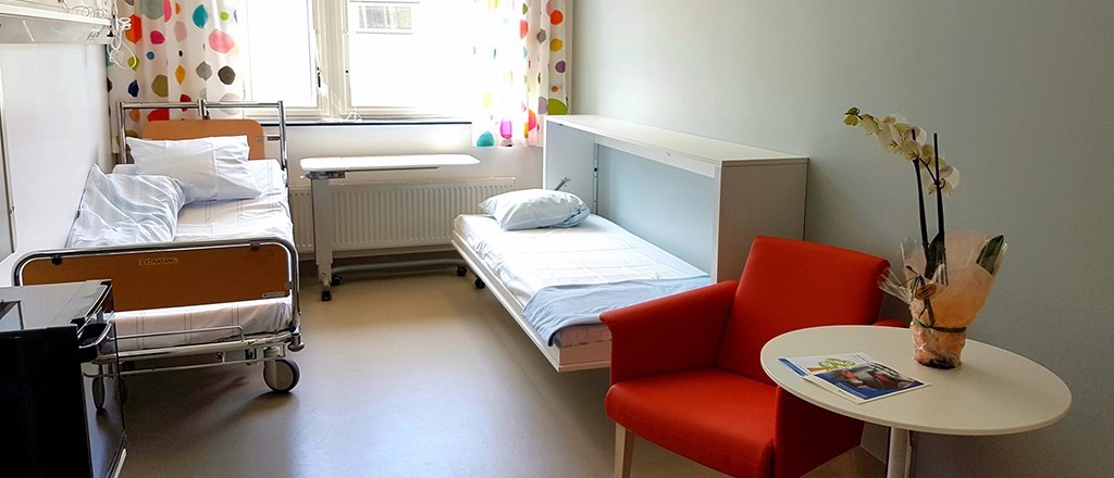 En sjukhussäng längs ena sidan av rummet och en utfällbar säng längs andra sidan. En fåtölj och ett litet bord. Fönster med stort ljusinsläpp.