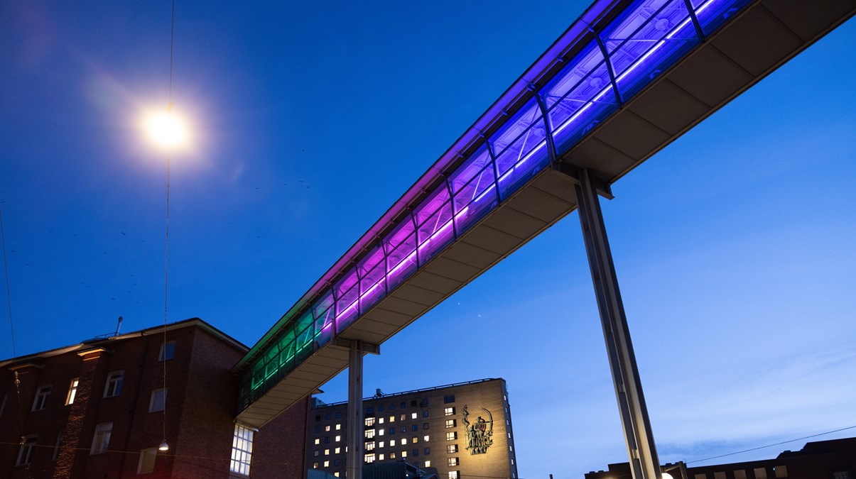Vid skymning upplyst gångbro vid Sahlgrenska sjukhuset. Bron lyser i färgerna grönt, rosa och blått.