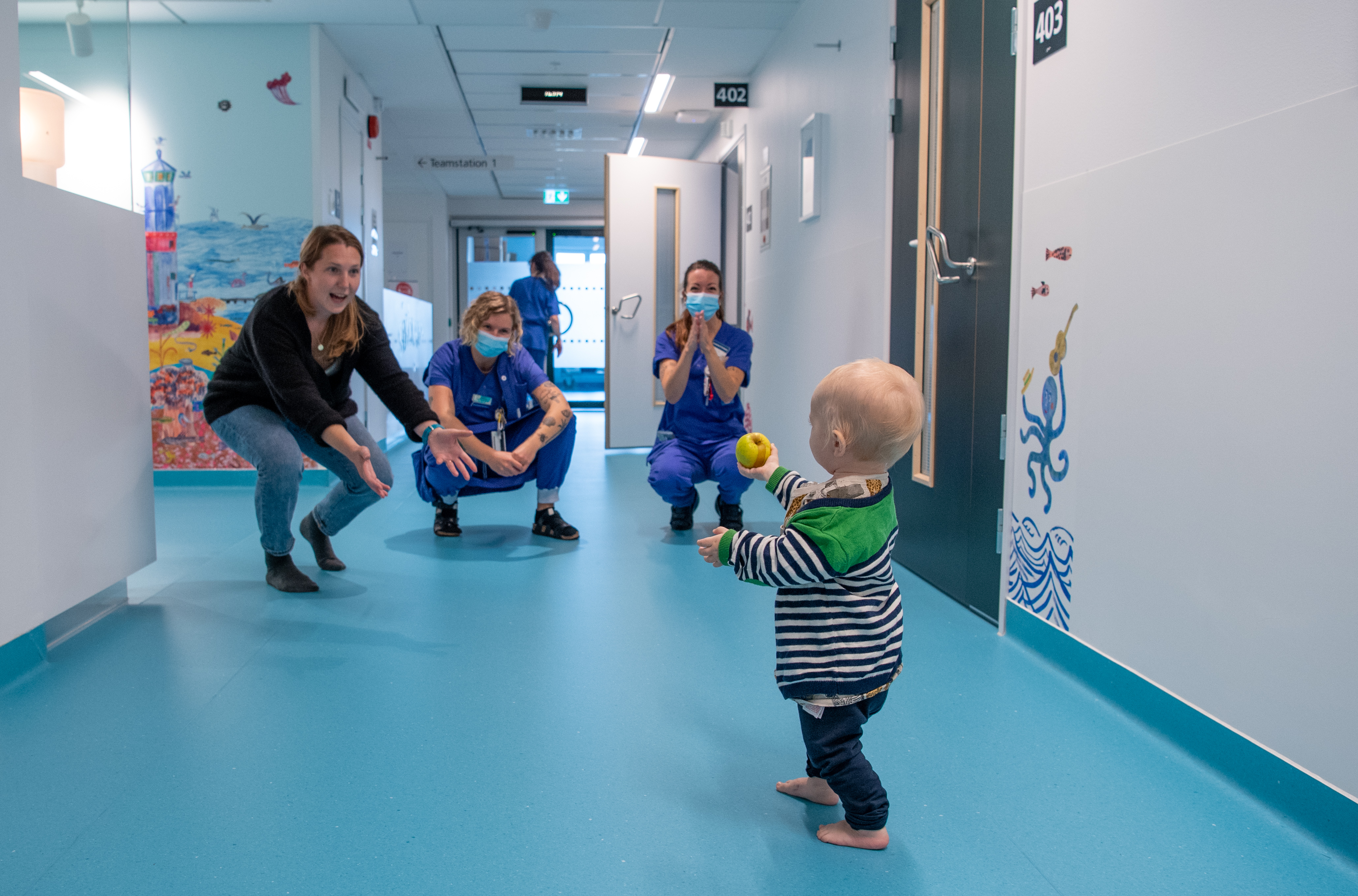 Litet barn med boll i vårdkorridor med blått golv sträcker ut en hand till sin mamma i svart tröja och jeans samt två i vårdpersonalen klädda i blåa scrubs