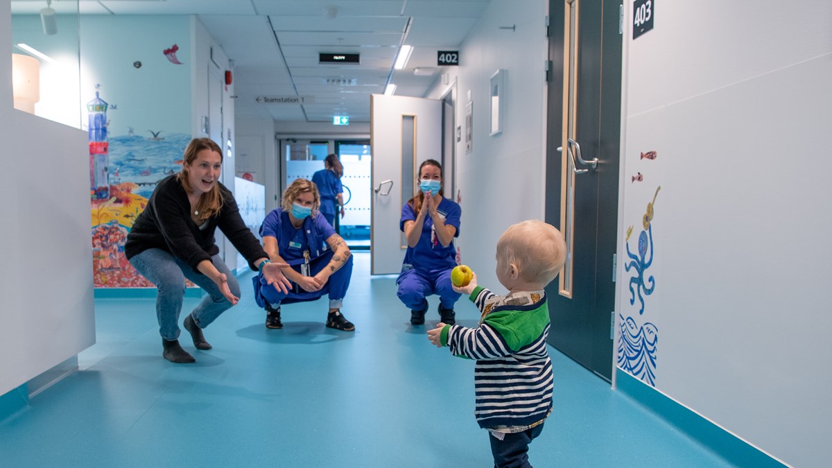 Litet barn med boll i vårdkorridor med blått golv sträcker ut en hand till sin mamma i svart tröja och jeans samt två i vårdpersonalen klädda i blåa scrubs
