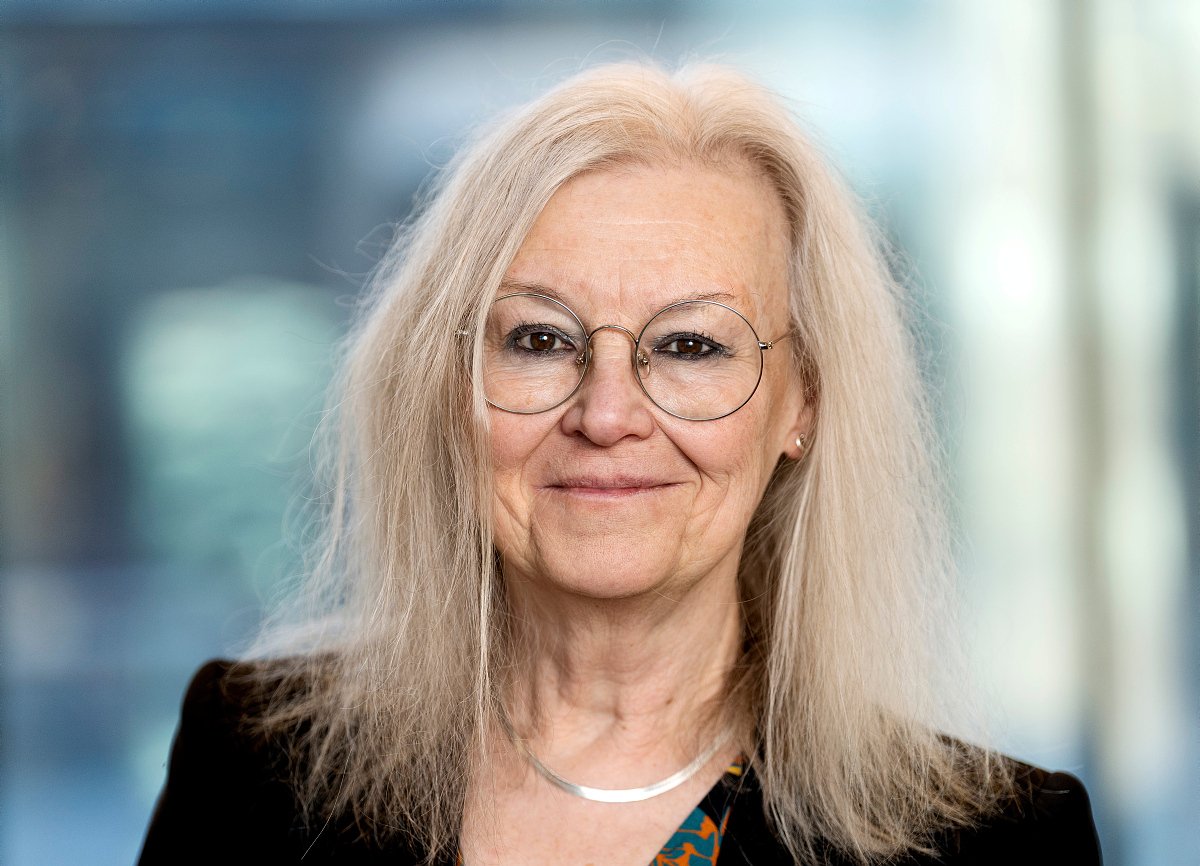 A woman with blonde hair and round glasses, smiling into the camera.