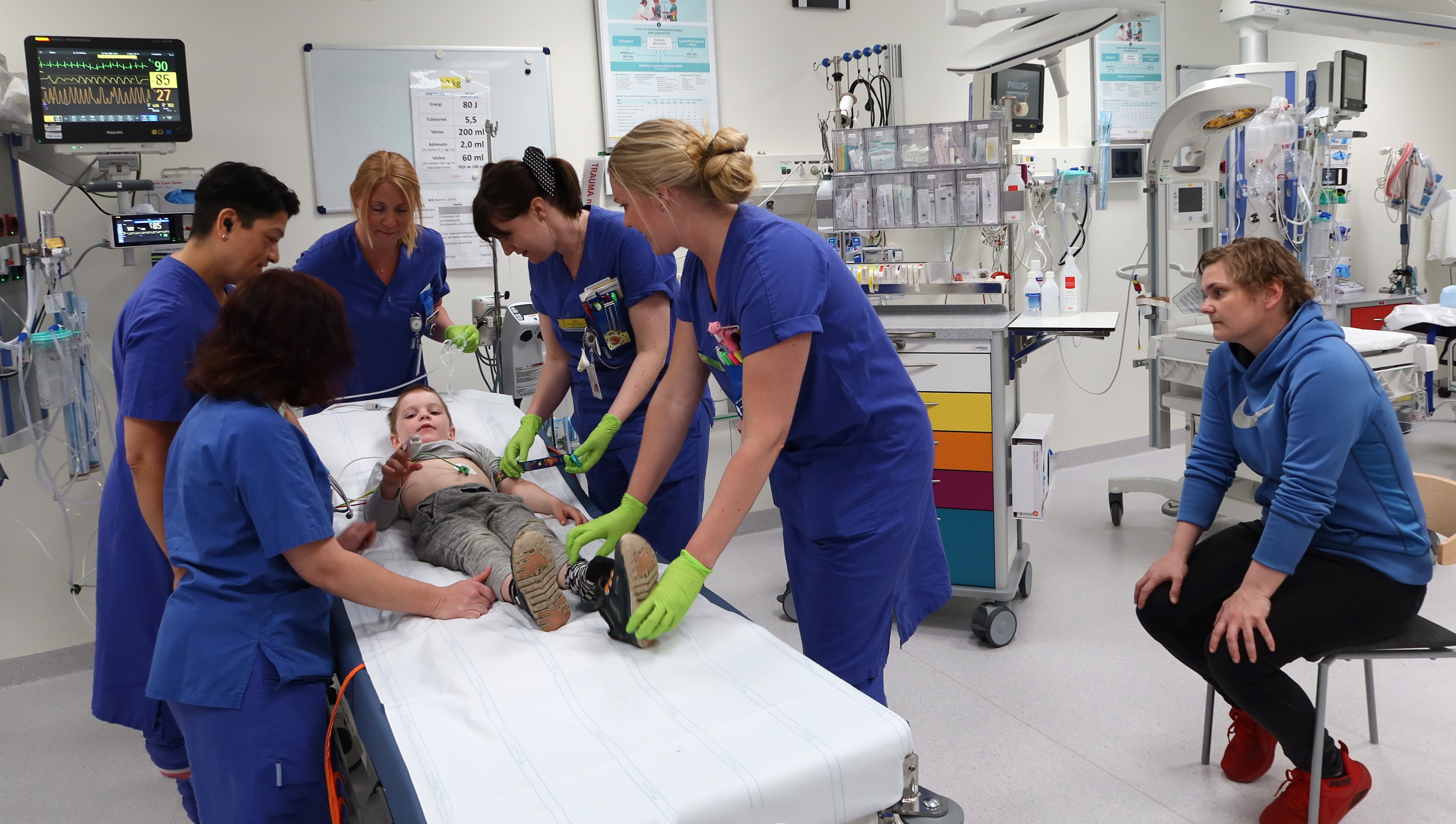 Ett barn ligger på en brits. Många människor står runtom.