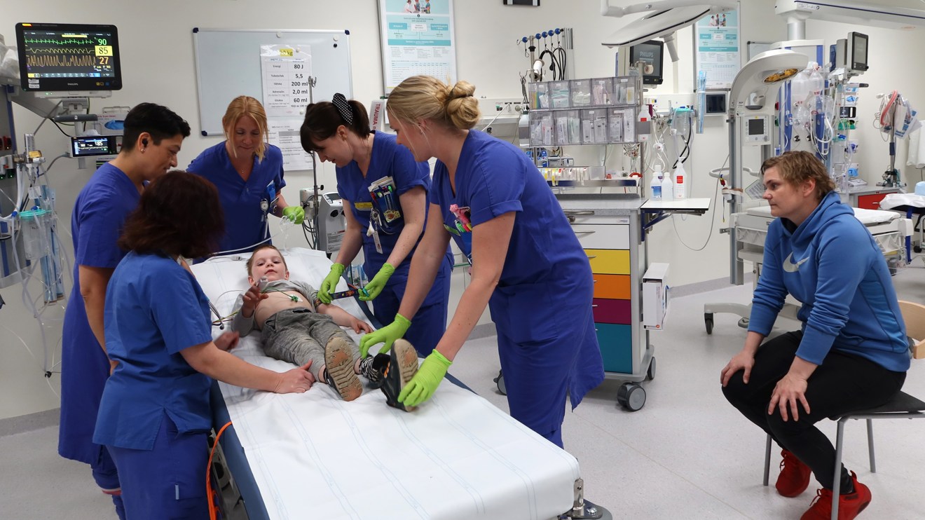 Ett barn ligger på en brits. Många människor står runtom.