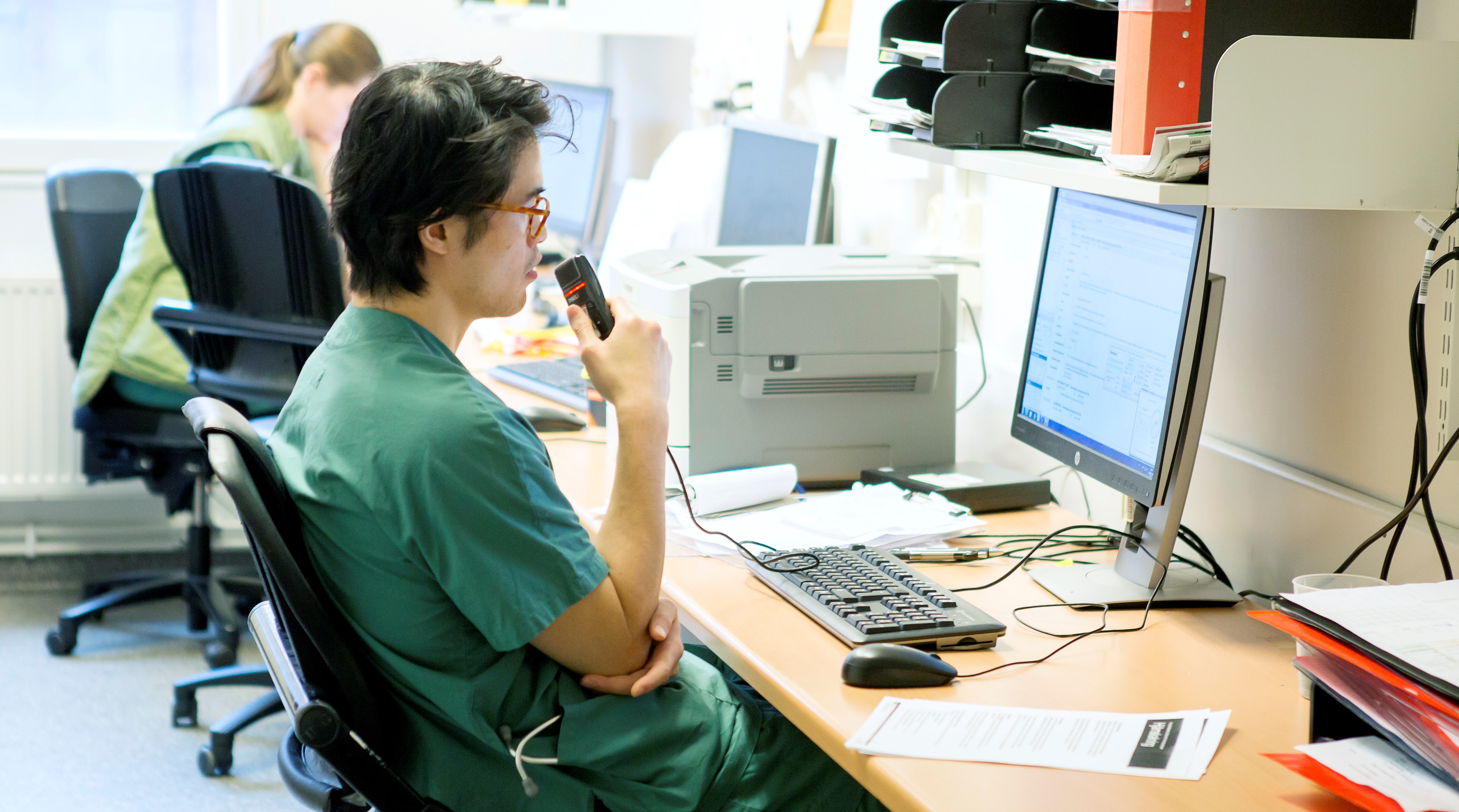 Läkare dikterar in i mikrofon vid skrivbord.