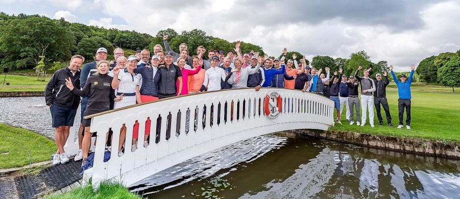 Deltagare i barngolften står på en liten bro över ett vattenhinder på golfbanan och ser glada ut.