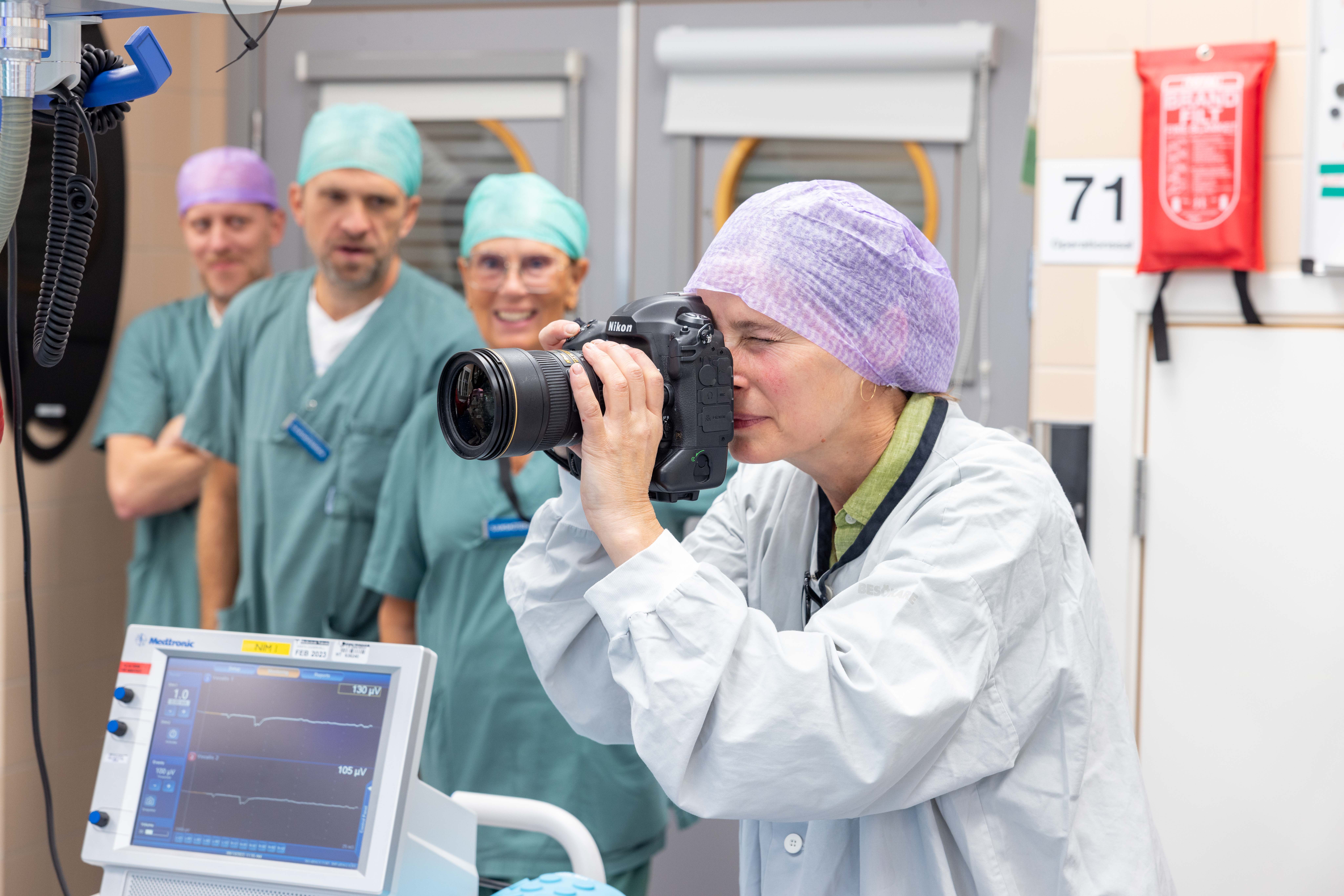 Kvinna fotografera i vårdmiljö. Tre läkare syns i bakgrunden.