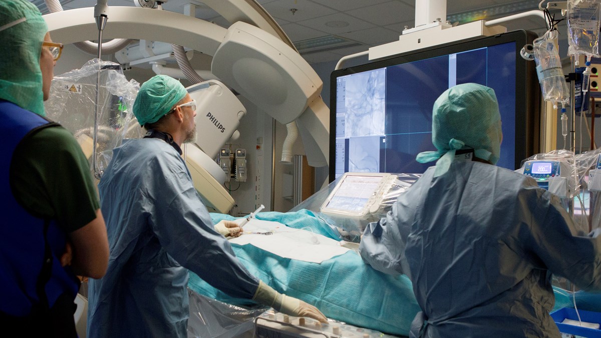 Tre medarbetare står och tittar på en stor skärm i en hybridoperationssal under en operation.