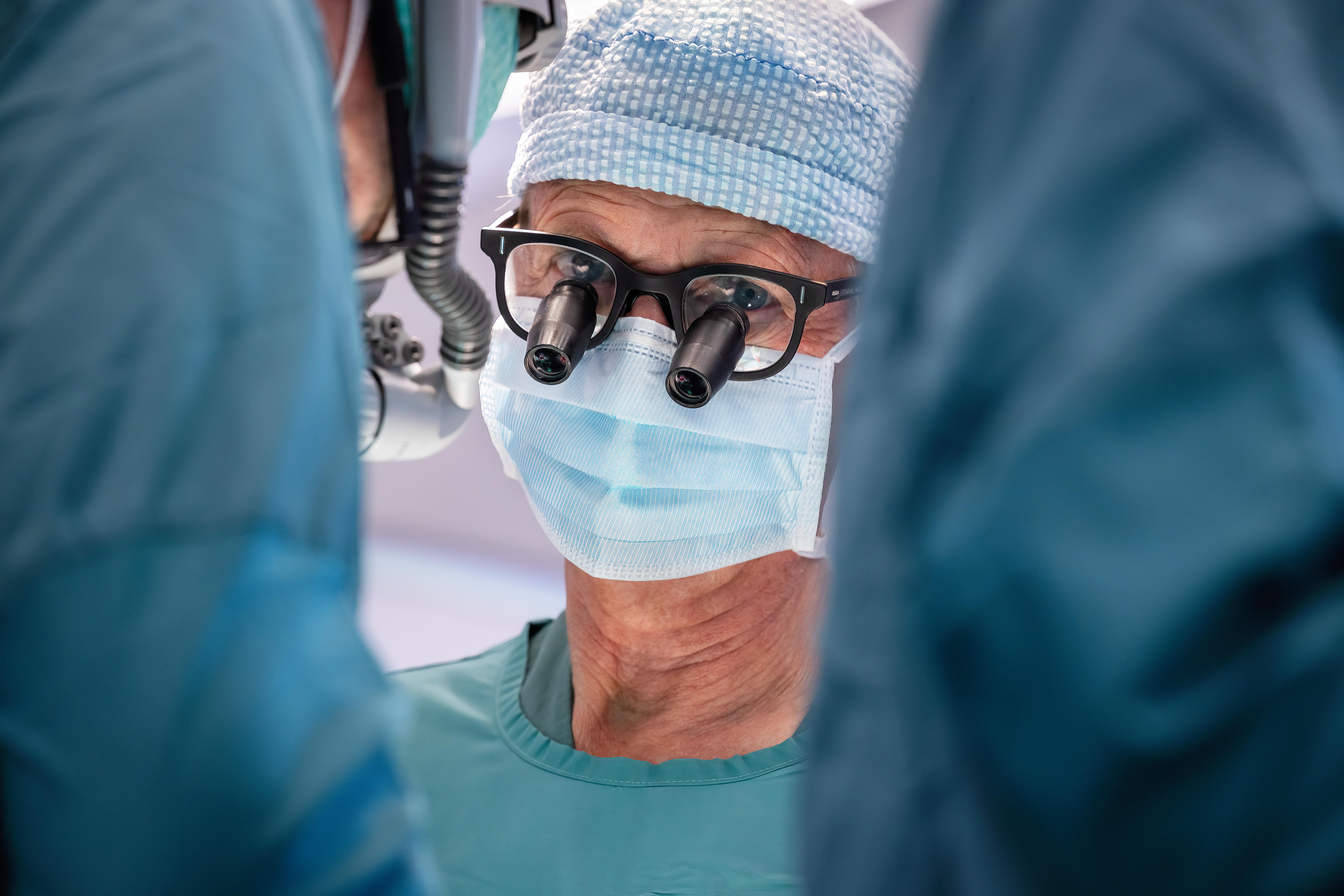 Male healthcare professional in surgical scrubs wearing magnifying loupes and a face mask.