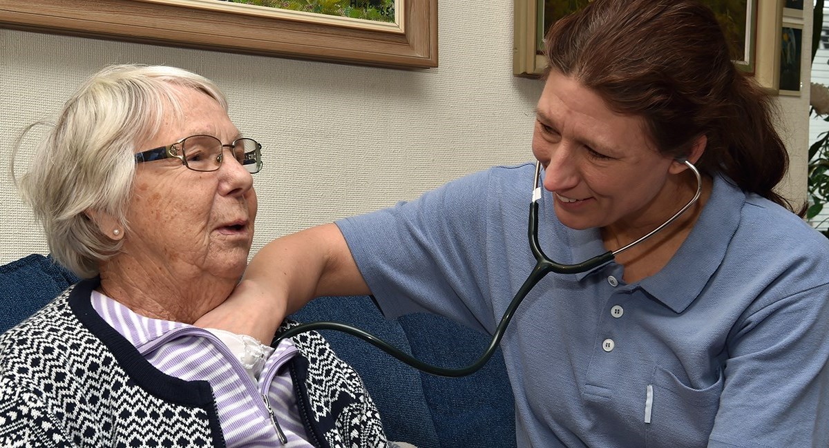 En läkare lyssnar på en äldre patients hjärta med hjälp av ett stetoskop. De sitter i en blå soffa hemma hos patienten.