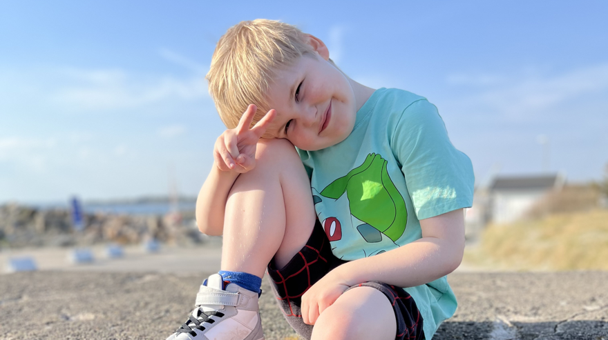 Pojke med blont hår iklädd t-shirt och shorts sitter på en klippa och ler mot kameran.