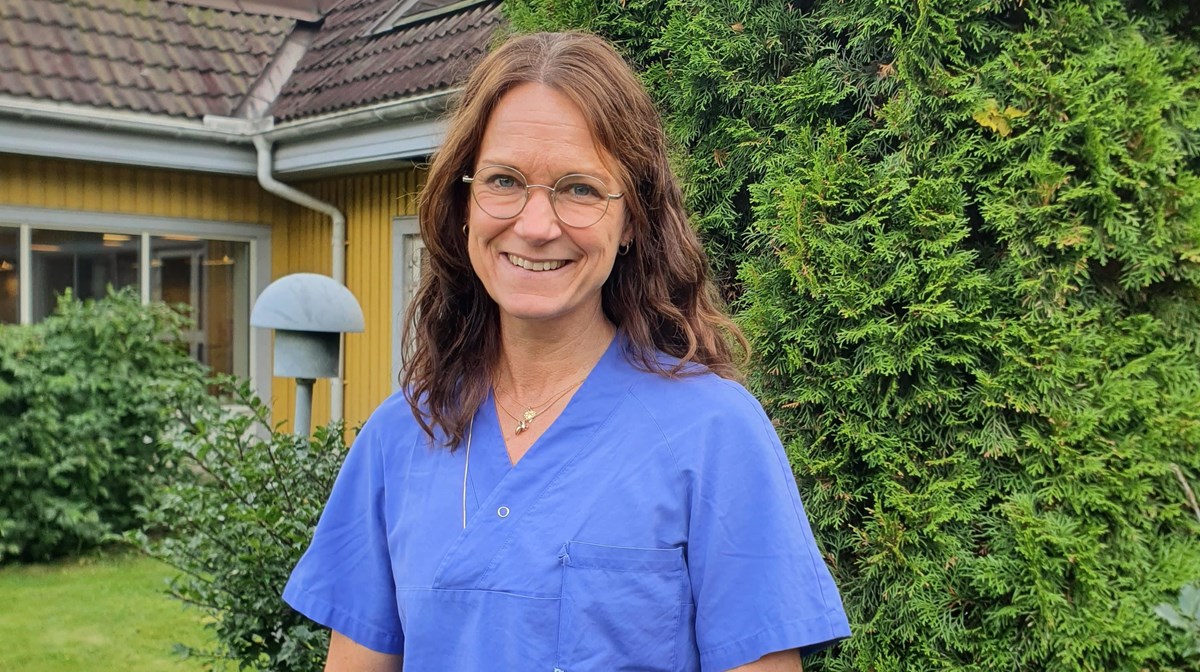 Leende Johanna Weichbrodt iklädd blå sjukhuskläder stående framför grön buske.