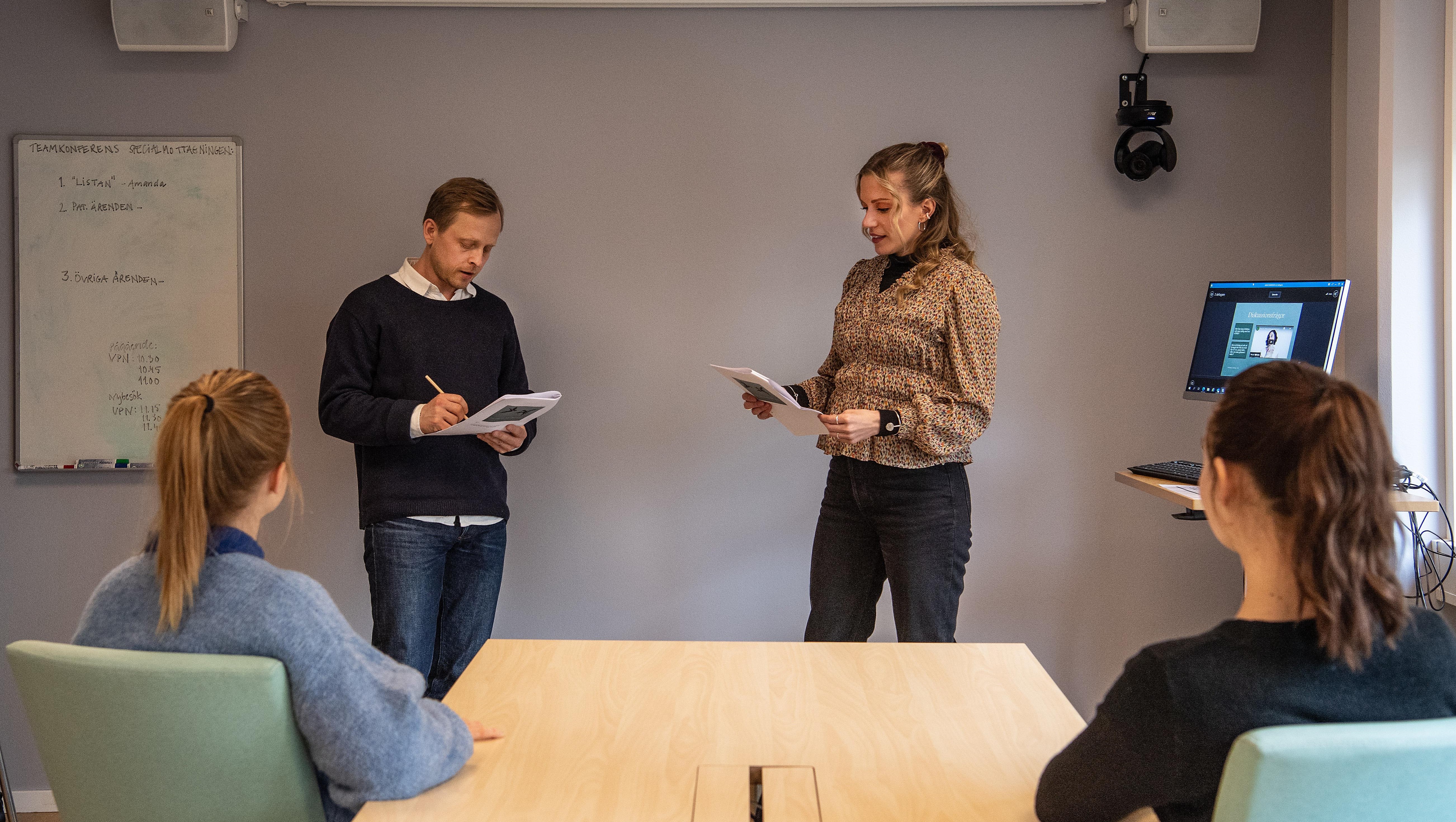 Två personer som står vi ett bord och pratar, två personer sitter och lyssnar