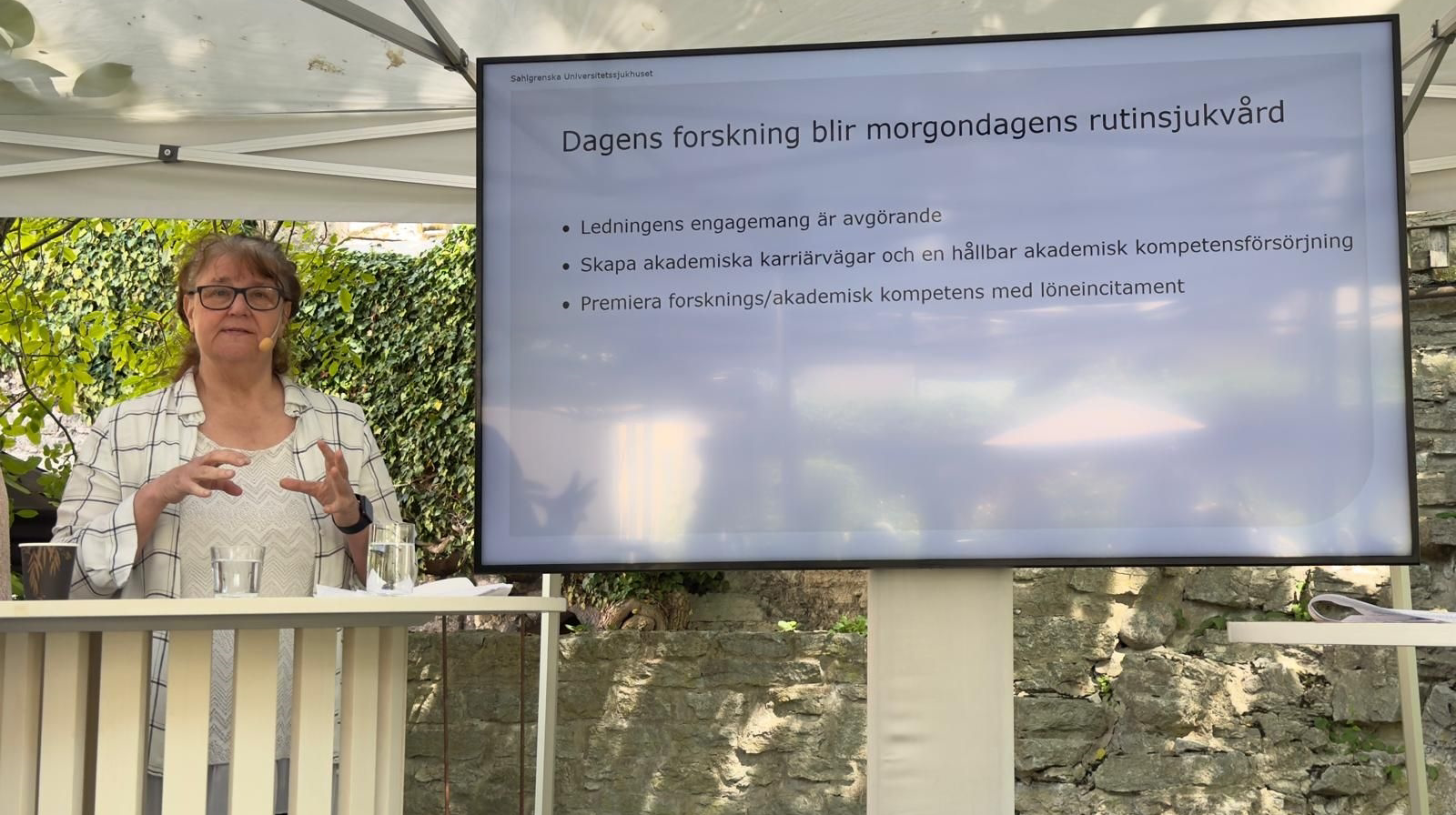 Kvinna framför en stor skärm på scen, utomhus under tak.