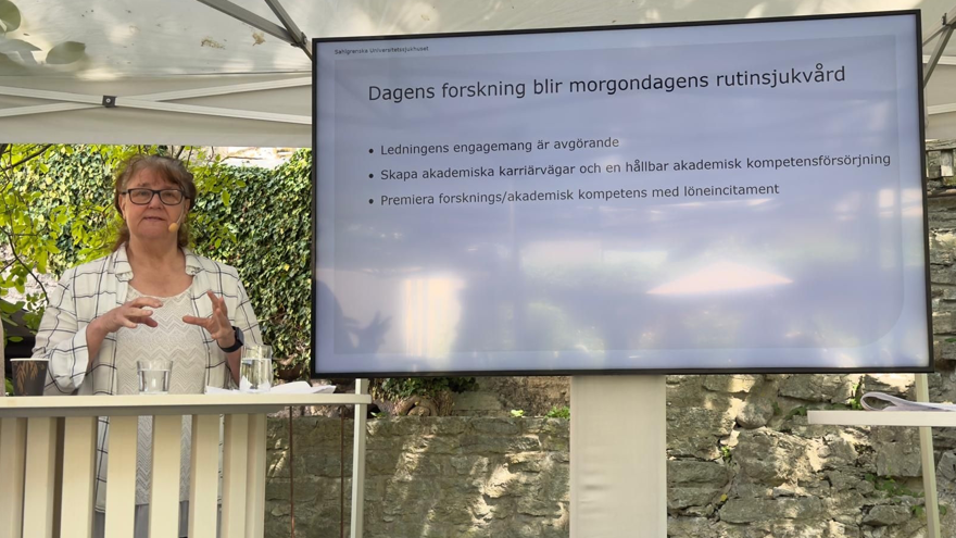 Kvinna framför en stor skärm på scen, utomhus under tak.