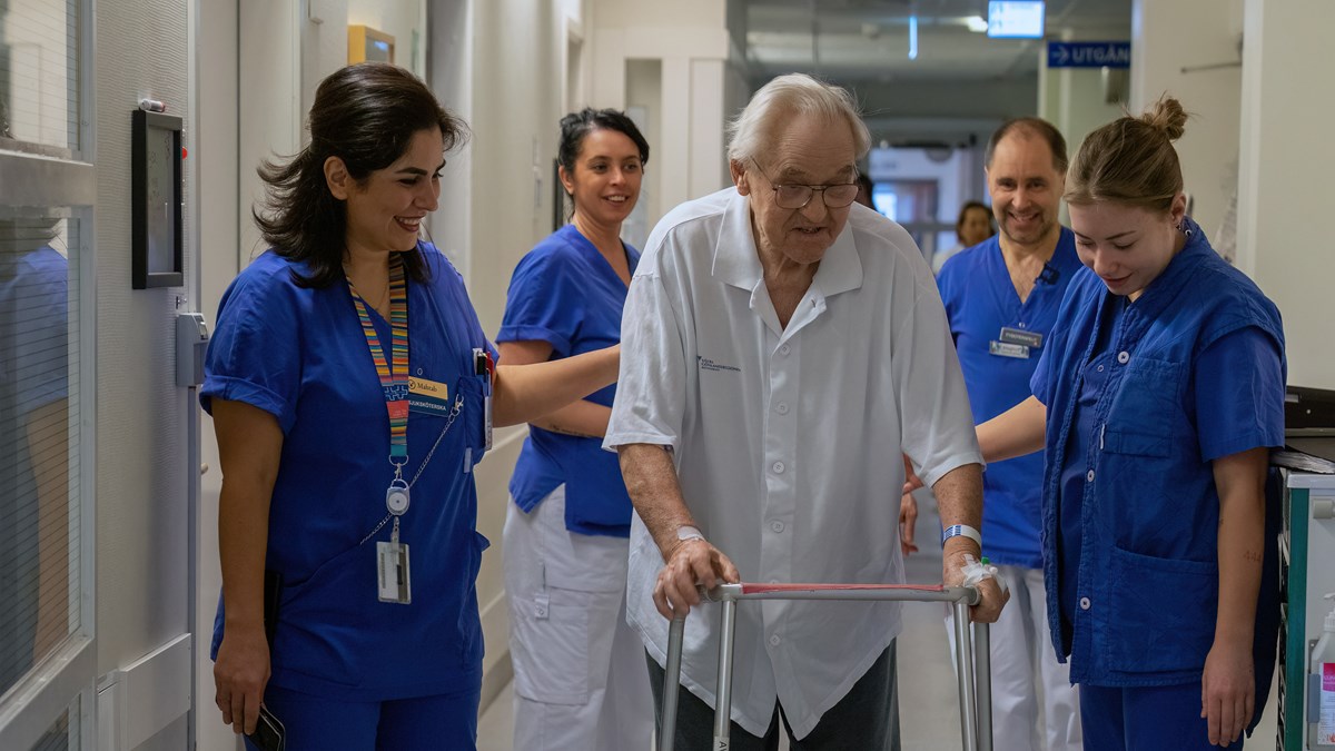 Äldre man med rullator i vita och gråa sjukhuskläder omringad av blåklädd vårdpersonal