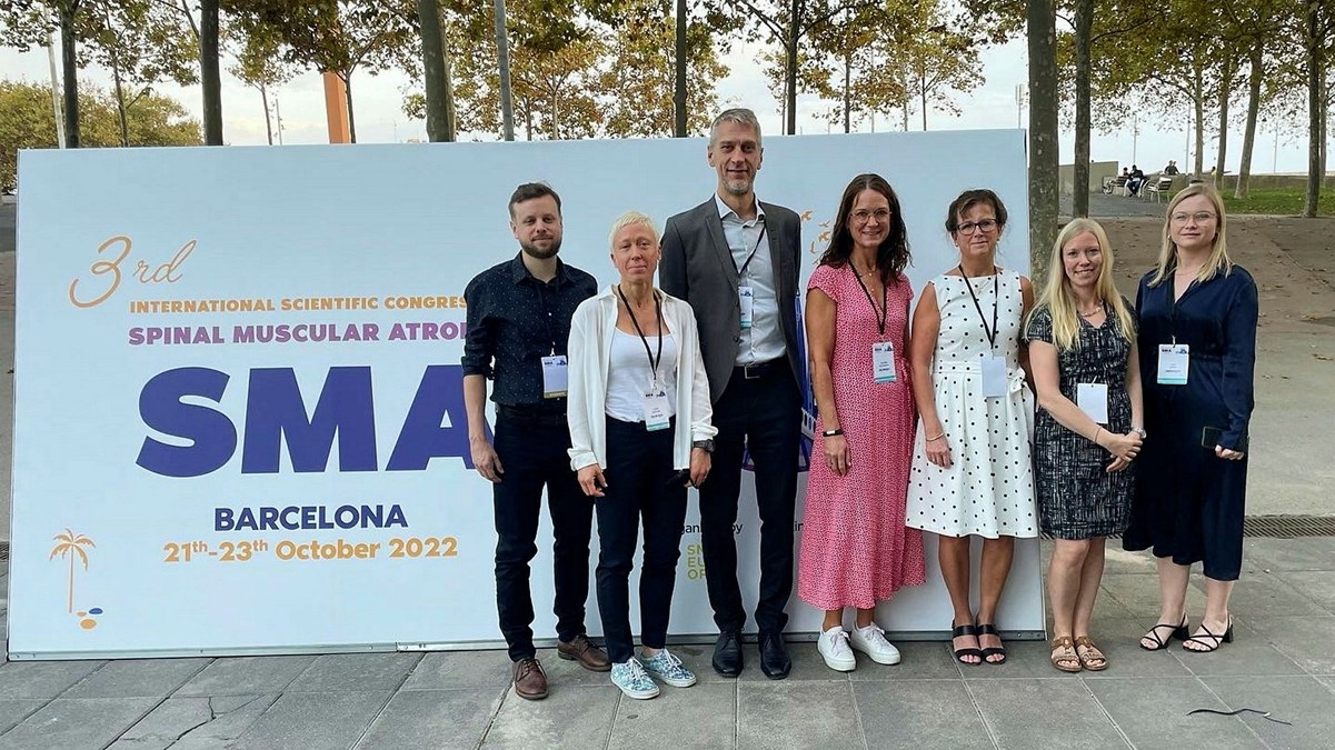 Det neuromuskulära teamet, står utomhus framför en skylt för en SMA-konferens i Barcelona.